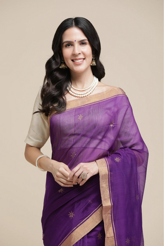 Woman wearing a purple saree with gold border on a beige background
