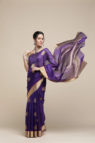 Woman wearing a purple saree with gold patterns against a beige background