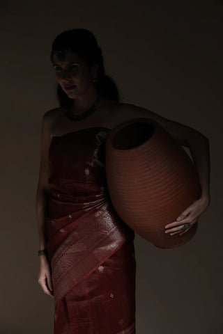 Woman in a red saree holding a large brown pot against a dark background