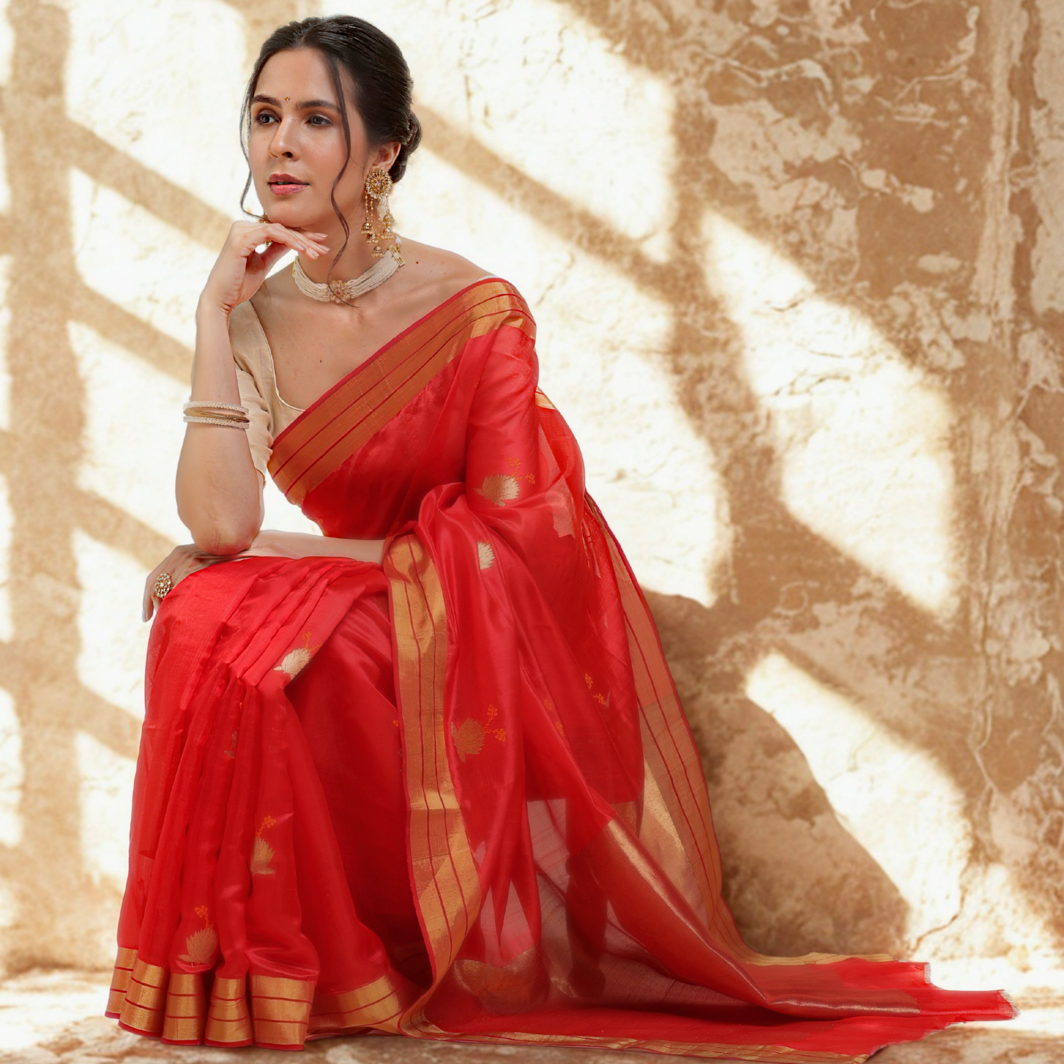 Woman in a red saree with gold accents sitting against a textured wall.