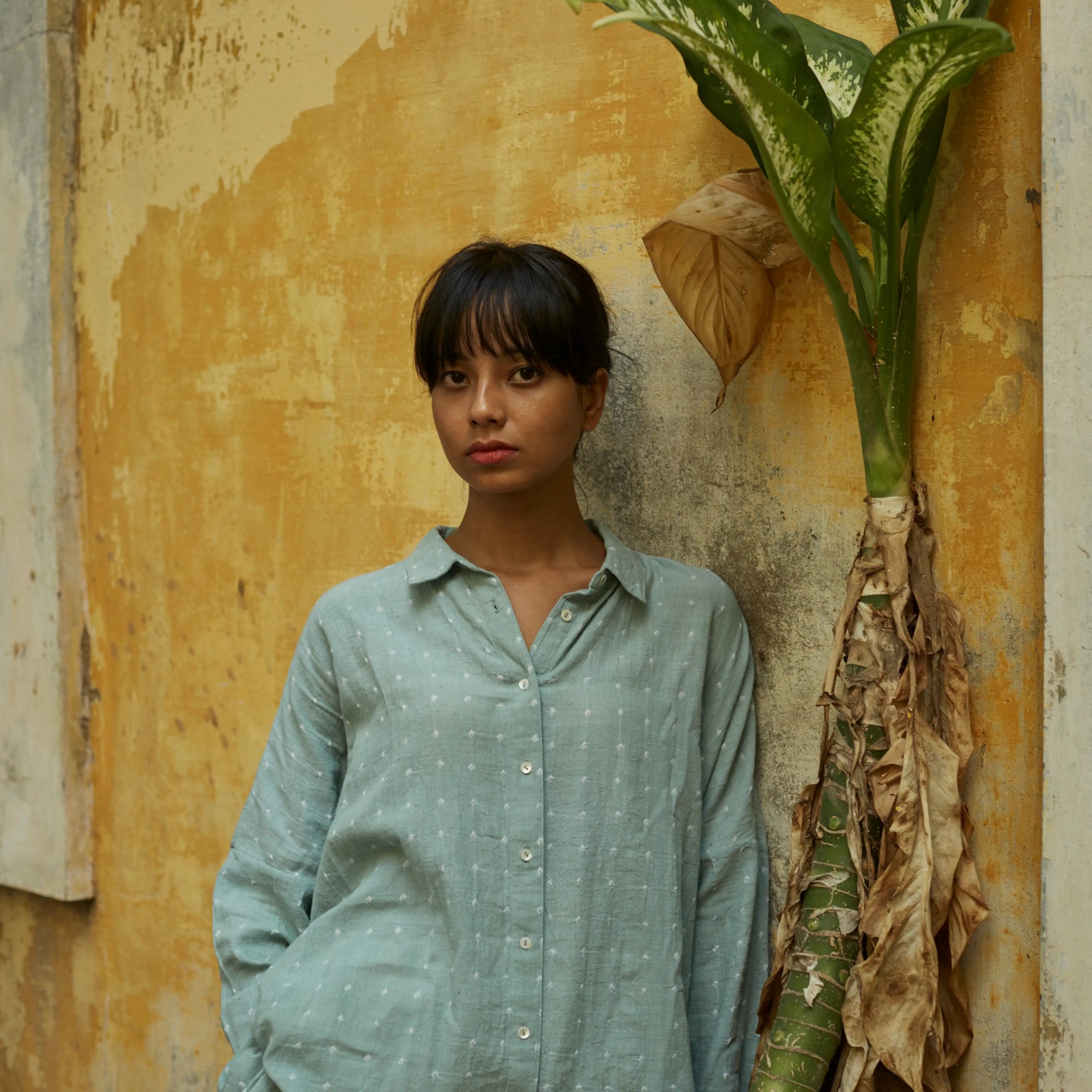 Woman in a light green shirt standing against a textured wall with a plant.